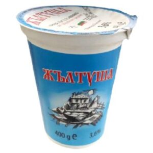 The image shows a container of yogurt with a blue background. The label features red Cyrillic text. Below the text is a black-and-white drawing of a rustic house with what appears to be a village or countryside setting. The container indicates that it contains 400 grams and has a 3.6% fat content. The top of the container also displays additional information, including a flag, possibly indicating the product's origin.