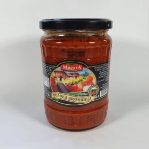 The image displays a jar of Bulgarian lutenitsa with a black lid. The label features a colorful illustration of fresh vegetables, including eggplant, chili peppers, tomatoes, and garlic, set against a country cottage background. The brand name appears prominently near the top, and there is additional text on the label likely describing the product, which is a traditional vegetable spread. The jar is sealed and appears to be full.