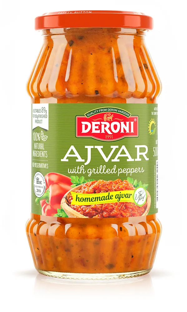 The image shows a jar of Deroni brand ajvar, which is a type of relish made with grilled peppers. The jar has a red lid and a label prominently featuring the brand name Deroni with the year 1991 below it. The label includes the phrase ajvar with grilled peppers in white text on a green background. There is an image of the ajvar with red peppers and greenery on the label, along with the text homemade ajvar. The jar's content is visible through the clear glass, showing a reddish-orange relish with specks of seasoning. The label also mentions 100% natural ingredients and no preservatives.