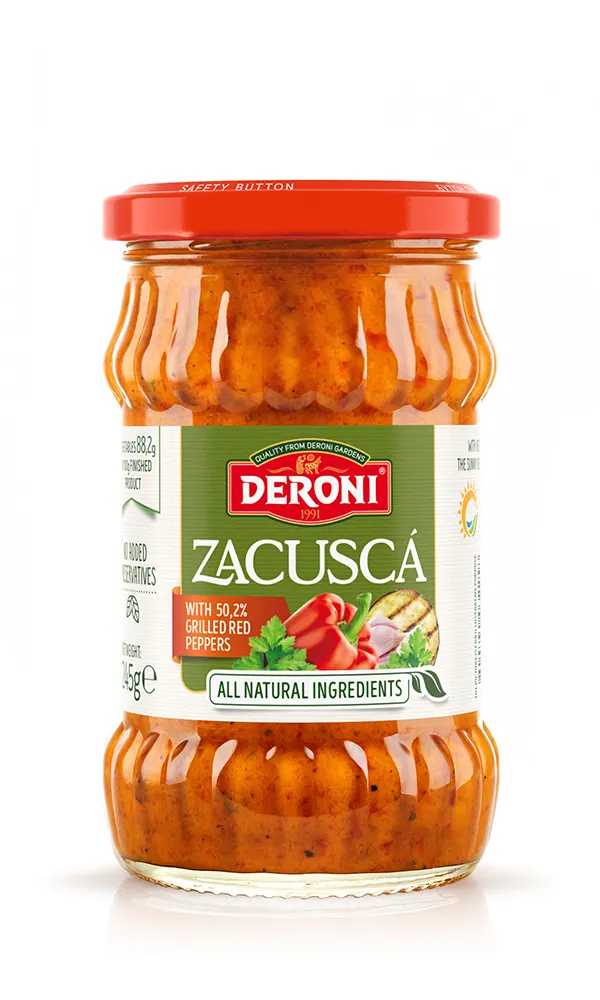 The image shows a jar of Deroni Zacuscă, a spread made with grilled red peppers. The jar is clear, allowing the viewer to see the chunky, reddish-orange spread inside. It features a red lid marked with Safety Button. The label on the jar highlights “All Natural Ingredients” and specifies 50.2% Grilled Red Peppers as a key ingredient. There are images of vegetables such as red peppers and garlic on the label, along with the Deroni brand logo. The net weight is indicated as 545 grams.