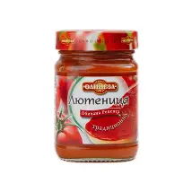 The image shows a jar of lyutenitsa, a traditional Balkan condiment. The label features a red and green color scheme, with images of tomatoes and possibly peppers, which are common ingredients. The text on the label appears to be in Cyrillic script, suggesting it is from Bulgaria or a surrounding region. The jar is sealed with a red lid.