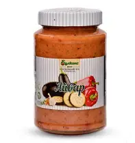 The image shows a jar of Ajvar with a white ribbed lid. The label features images of an eggplant, a red bell pepper, and garlic. The name Ajvar is prominently displayed, suggesting it's a type of pepper and eggplant spread. The jar has a clear view of the reddish-orange spread inside.