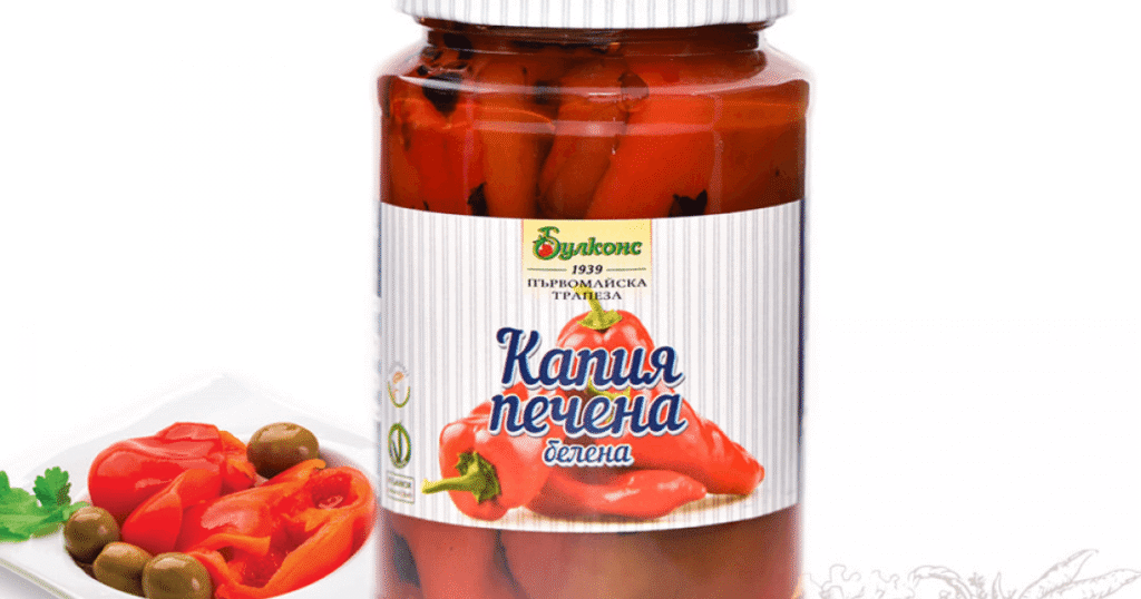 The image features a jar labeled in Bulgarian containing roasted and peeled red kapia peppers. The jar has a white lid and a label with an image of the peppers. To the left of the jar, there is a small plate with roasted peppers, green olives, and a sprig of parsley.