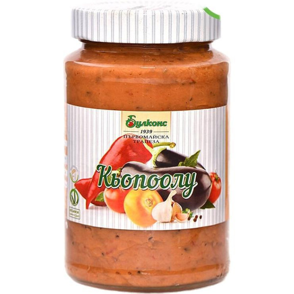 The image shows a jar of Kyopoolu, a type of vegetable spread. The label has images of red peppers, eggplants, tomatoes, and garlic, suggesting the ingredients. The brand name appears at the top of the label, and the lid has a striped design. The text on the label is in a Cyrillic script.