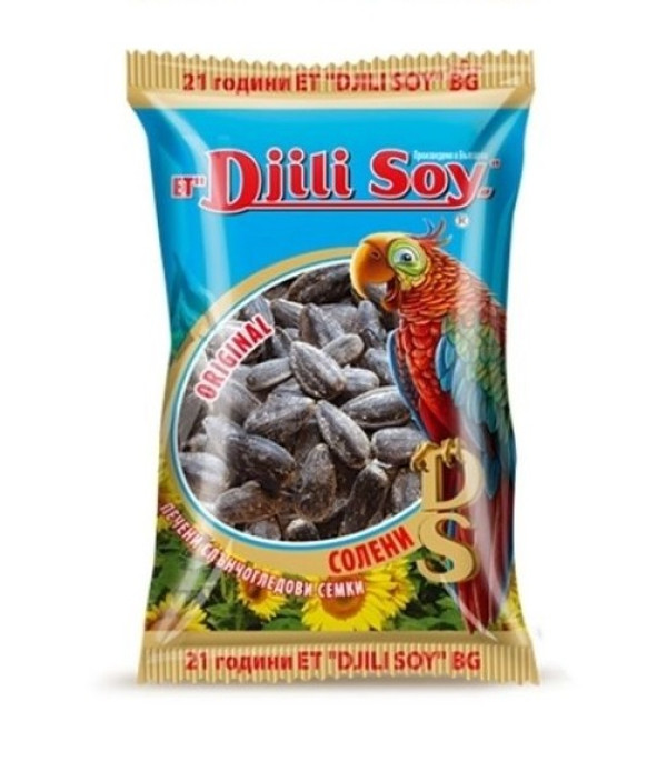 The image shows a package of Djili Soy sunflower seeds. The packaging is predominantly blue with a colorful parrot illustration on the right side. The center features a transparent circular window showing the seeds inside and text indicating ORIGINAL. There are sunflower illustrations at the bottom, and various text elements, presumably in Bulgarian, including Солени (salty) and Д печени слънчогледови семки (roasted sunflower seeds). The package top edge reads 21 години ET D.JILI SOY BG.