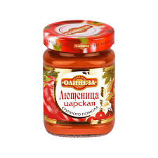 The image shows a glass jar with a red lid, containing a spread or sauce. The label on the jar features Cyrillic text and visual elements like red peppers and tomatoes, indicating it is a type of vegetable spread. The brand appears to be Олинеза.