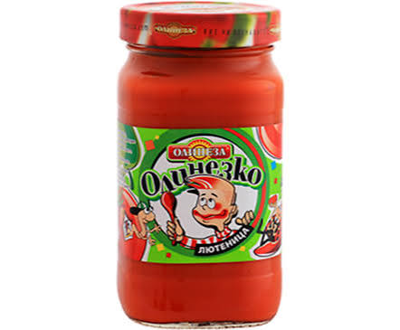 The image shows a jar containing a red sauce, likely a type of lutenitsa. The label on the jar is colorful and features a cartoon character, which appears to be a smiling chef holding a spoon. The brand name is written in bold letters, and the design includes various bright colors, mainly green and red. The lid of the jar is also red.