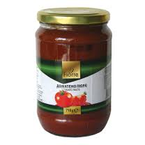The image shows a glass jar of tomato paste. The jar has a gold lid and a label with a green and white design. There are images of tomatoes on the label, along with text. The brand name appears to be Fiore, and there is a weight indication of 710g.