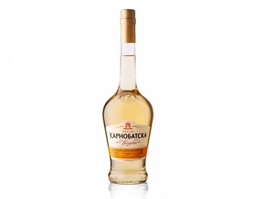 The image shows a bottle of KARNOBATSKA rakia. The bottle is clear glass with a distinct, rounded shape and has a long neck. The rakia is a light amber color. The label on the front features text in Cyrillic and includes a logo with a coat of arms. There's additional text on an orange band at the bottom of the label. The cap is golden in color. The bottle is set against a plain white background.