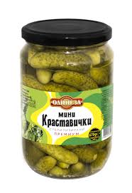 The image shows a glass jar filled with pickled mini cucumbers. The label on the jar has a predominantly green and yellow color scheme, with Cyrillic text indicating it's a product of pickled mini cucumbers. The jar has a black lid.