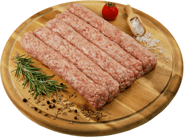 The image shows a wooden circular board containing five uncooked, cylindrical minced meat sticks arranged in parallel. A small tomato lies at the top, and to the right, a wooden scoop holds coarse salt. Sprigs of rosemary and scattered spices, including peppercorns and caraway seeds, are placed around the meat on the board.