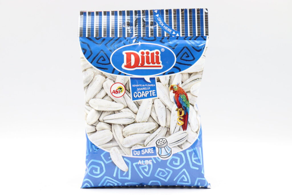 The image depicts a packaged product labeled Dijili, featuring a design with sunflower seeds prominently displayed. The packaging is primarily blue with decorative geometric patterns. There's a colorful parrot illustration on the front. The text indicates the product is roasted sunflower seeds (semințe de floarea soarelui coapte) with salt (cu sare). The top of the package has a striped design with an Open Here label.