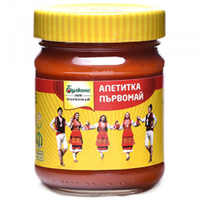 The image shows a jar of Апетитка Първомай sauce. The jar has a yellow lid and label. The label features a group of people in traditional clothing, holding hands, and dancing. The background of the label is yellow with red accents, and there are Cyrillic characters, denoting the product name and brand.