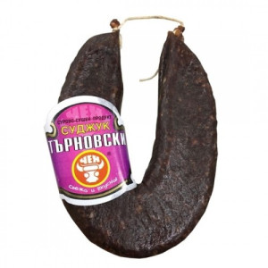 The image shows a looped, dark-colored sausage, likely cured or smoked, as it has a firm, dry texture. It is held together with a string at the top. There is a label attached to it that displays some Cyrillic text in white and purple colors, along with a logo or symbol at the bottom. The overall appearance suggests it's a type of traditional or specialty sausage.